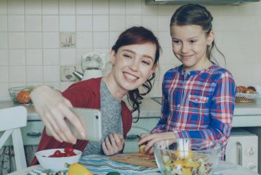 Smiling mother making selfie portrait together with young cute daughter cooking breakfast at home in kitchen. Family, cook, and people concept